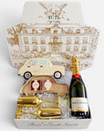 A luxury gift set from Yuti, featuring a bottle of Moët & Chandon champagne, gold-finished champagne flutes and a car-shaped box, all arranged inside a white gift box with a gold-toned illustration of a grand building on the lid.