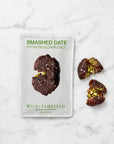 A package of Smashed Date Pistachio & Dark Choc Wild+Tempered chocolate dates on a white marble background, with two broken dates showing their pistachio filling next to the package.