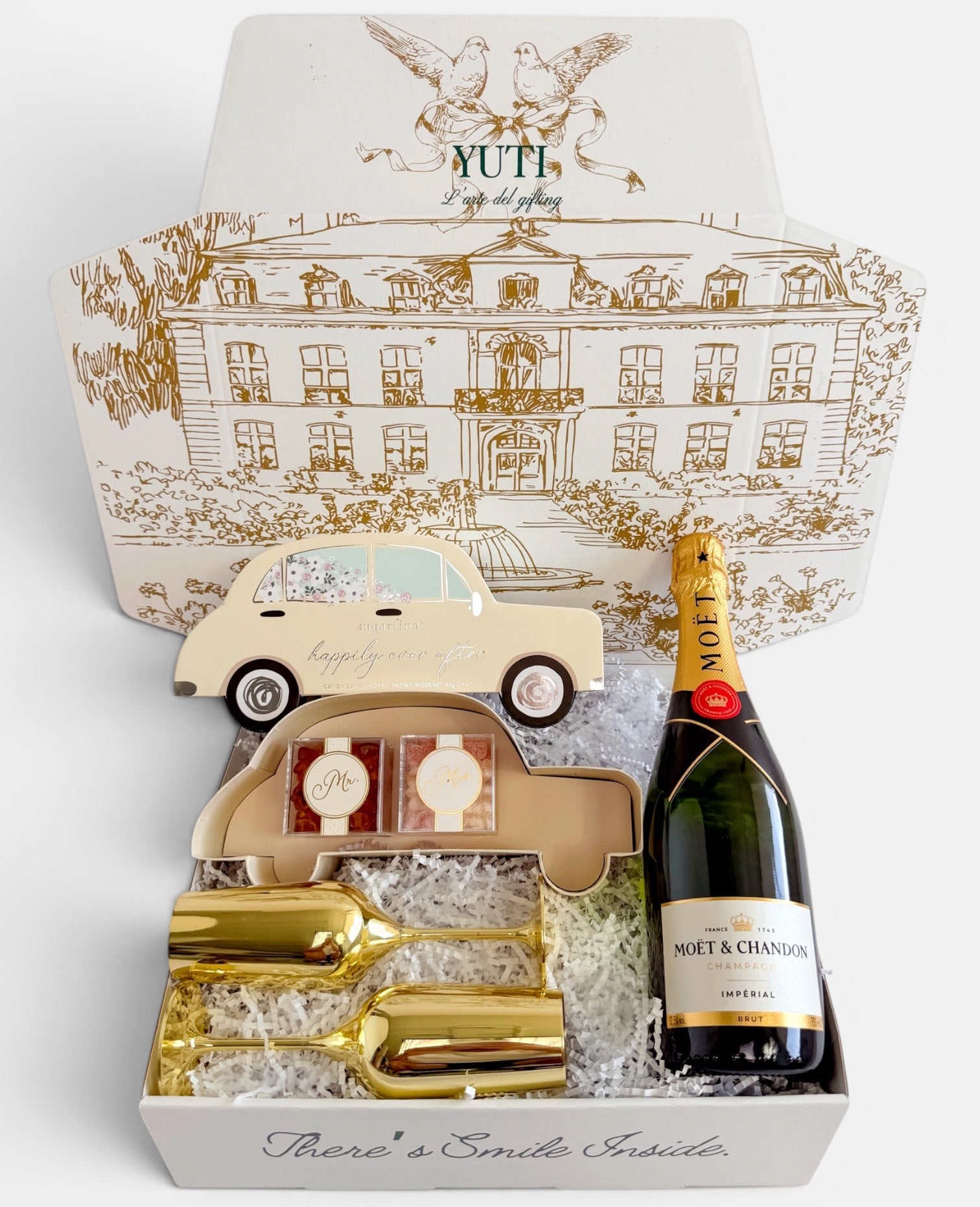 A luxury gift set from Yuti, featuring a bottle of Moët &amp; Chandon champagne, gold-finished champagne flutes and a car-shaped box, all arranged inside a white gift box with a gold-toned illustration of a grand building on the lid.