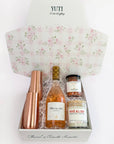 A curated gift box set by Yuti containing a bottle of Rosé Champagne, a rose gold cocktail shaker, a jar of rose petals, and a candle, all arranged inside a white box with a pink floral patterned lid.