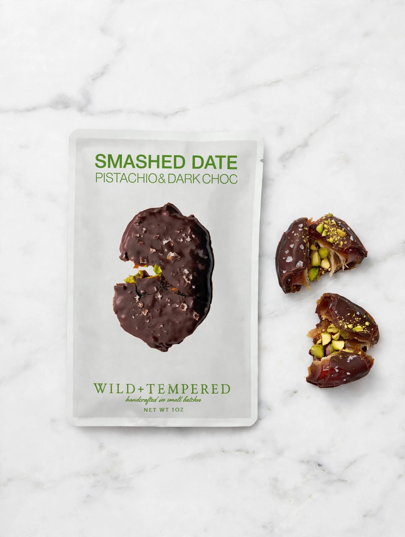 A package of Smashed Date Pistachio &amp; Dark Choc Wild+Tempered chocolate dates on a white marble background, with two broken dates showing their pistachio filling next to the package.